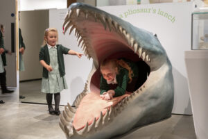 Two children playing with an exhibit of a large animals mouth. One girl is inside the mouth while the other is posed beside it