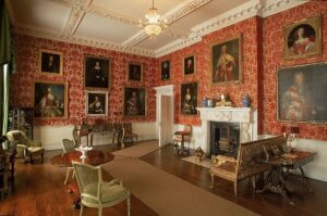 Interior of the Palladian style drawing room of Lydiard House - red flocked wallpaper and paintings depicting portraits. The room has chairs and tables and a large fireplace.